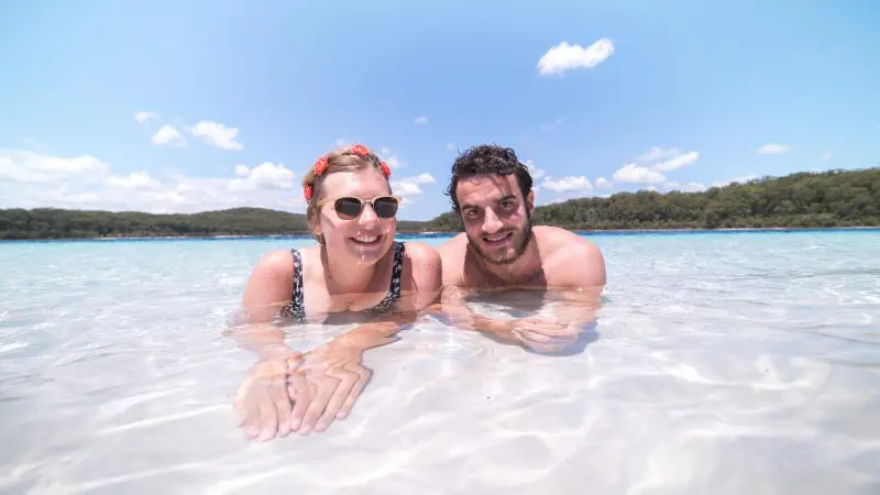 Happy couple wading in crystal-clear, shallow waters on a sunlit beach during Kgari Day Tour from Noosa, perfect for adventure.