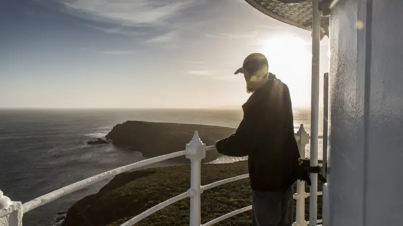 Traveller on Cape Bruny Lighthouse balcony at sunset enjoys panoramic ocean views during top-rated Bruny Island sightseeing tour.
