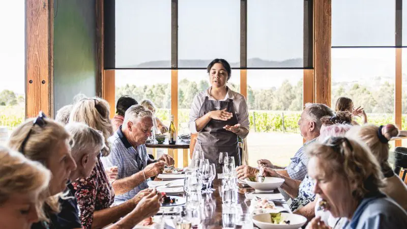 A lively group enjoys fine dining at a long table as a guide shares insights, evoking a Coal River Valley Wine & Gin Safari tour.