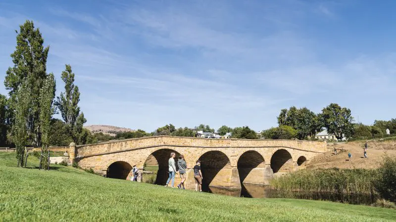 Visitors stroll beside a historic stone bridge on a Coal River Valley Wine and Gin Safaris tour, surrounded by lush trees and green grass.