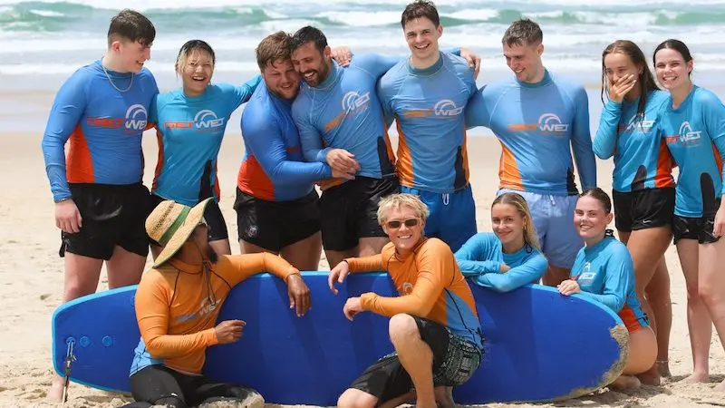 Group of surfers in blue rash vests posing with surfboards for a 2-hour beginner surf session, ages 13+, smiling on the beach.