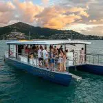 Experience a Sundowner Sunset Cruise on a floating boat bar as guests relax, with scenic mountains and shoreline views at golden sundown.