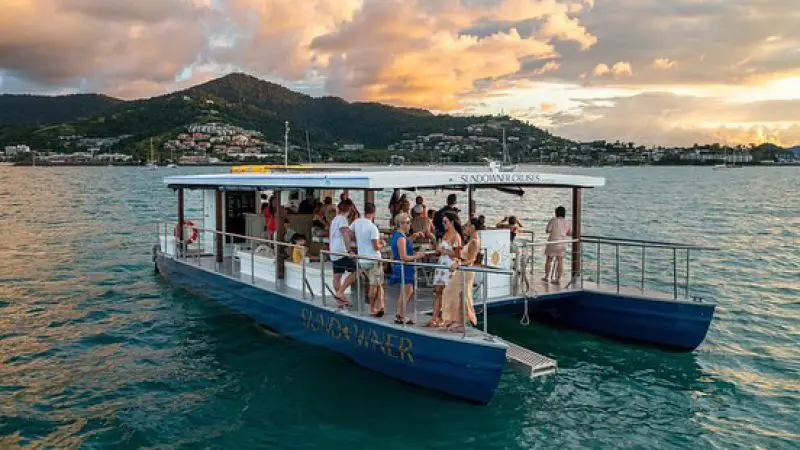 Experience a Sundowner Sunset Cruise on a floating boat bar as guests relax, with scenic mountains and shoreline views at golden sundown.