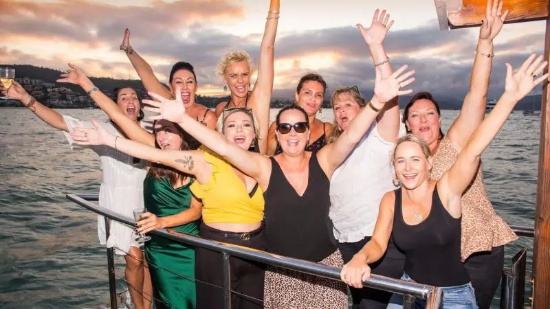 Smiling women celebrate on a Sundowner Sunset Cruise, raising arms with joy against a scenic water backdrop at golden hour.
