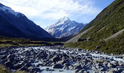 Scenic rocky river winds through lush green valley with snow-capped Mount Cook under clear blue sky, ideal for Queenstown tours.