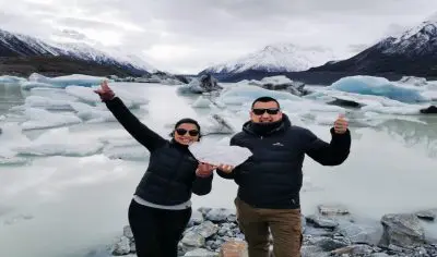 Two people in winter jackets pose and smile by sparkling ice at a snowy, mountain-framed glacial lake beneath New Zealand’s Mt Cook.