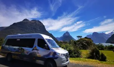 Scenic campervan by lush greenery, overlooking Queenstown’s mountains and lake under a vibrant blue sky with wispy white clouds.