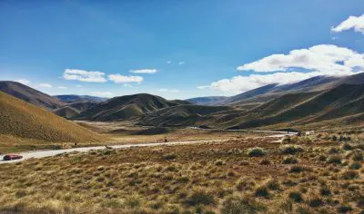Scenic winding road through golden hills beneath clear blue skies, with a red car driving between Queenstown and Mt Cook, New Zealand.
