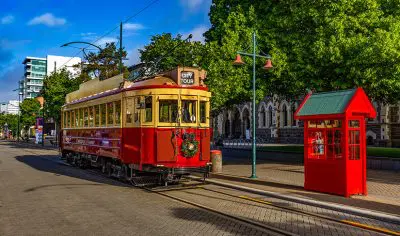 Vintage red tram and classic phone box on a sunlit Christchurch street, framed by leafy trees and iconic historic architecture.