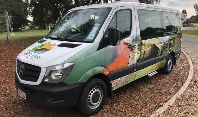 White and green nature-themed tour van parked on a picturesque, leaf-strewn roadside by Lake Tekapo with scenic views.