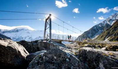 Tourists walk across a suspension bridge spanning rocky terrain, with snow-capped Mt Cook mountains and a clear blue sky overhead.
