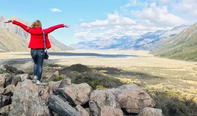 Adventurous woman in vibrant red jacket stands on rocky outcrop with arms wide, enjoying breathtaking Mt Cook valley and mountain views.