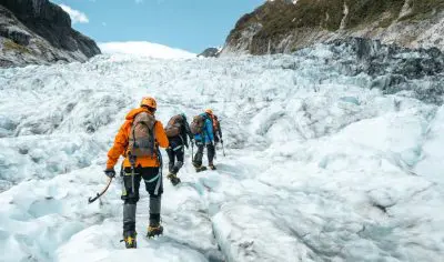 Three climbers in bright orange jackets trek up the snowy Franz Josef Glacier, framed by rugged mountains and a clear blue sky.