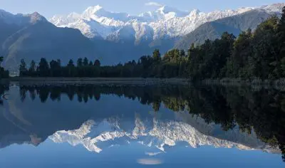 Breathtaking snow-capped peaks mirrored in a serene lake on a Wanaka to Franz Josef small group tour beneath a vibrant blue sky.