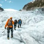 Three adventurers in colourful jackets trek across a pristine snowy glacier on a Wanaka to Franz Josef guided tour beneath blue skies.