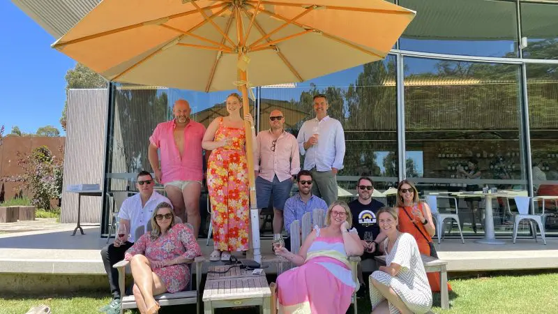 Thirteen happy guests smile together beneath a large umbrella during a sunny Yarra Valley Winery Tour with Laid Back Social.