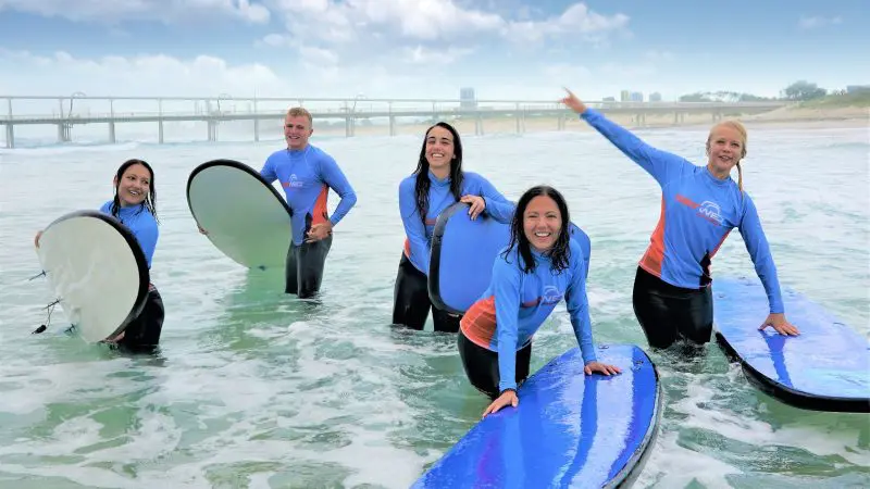 Five happy people in wetsuits enjoy a 2-hour beginner surf session for ages 13+ beside a scenic pier under cloudy skies.