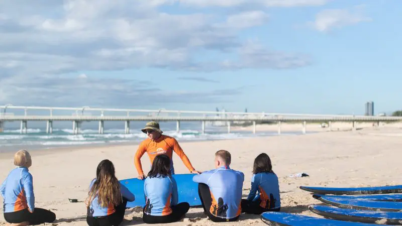 Surf instructor teaches five teenagers, aged 13 and over, during a 2-hour beginner surf lesson on a sunny beach with waves in the background.