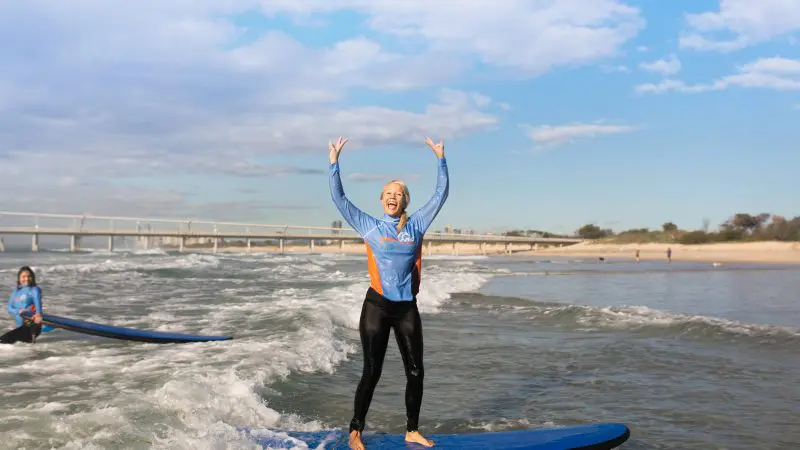 Participant smiles on a surfboard during a 2-hour beginner surf lesson, with scenic beach and iconic bridge in the vibrant background.