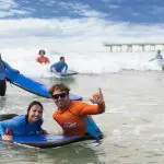 Teens and adults, 13+, grinning on a surfboard by the pier during a 2-hour beginner surf lesson—perfect for first-time surfers.