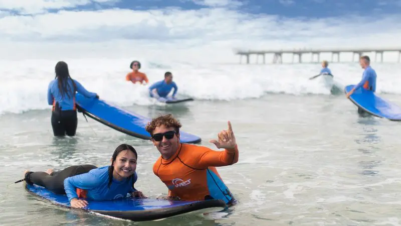 Teens and adults, 13+, grinning on a surfboard by the pier during a 2-hour beginner surf lesson—perfect for first-time surfers.