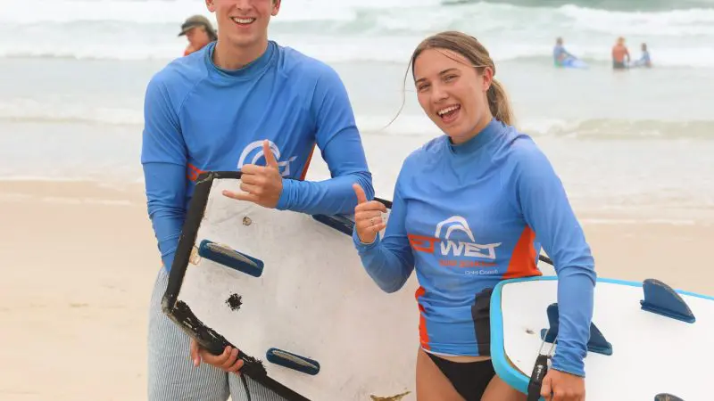 Two happy teenagers, aged 13+, smiling and giving a thumbs up after a beginner surf lesson on a sunny sandy beach, sea in view.