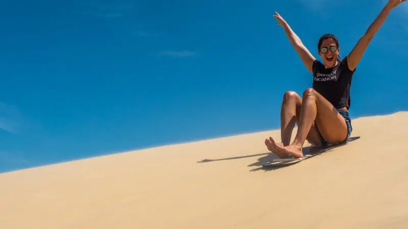 Smiling woman sandboards down golden dune with arms raised during a thrilling 1 Day Port Stephens Colourful Collective Travel tour.