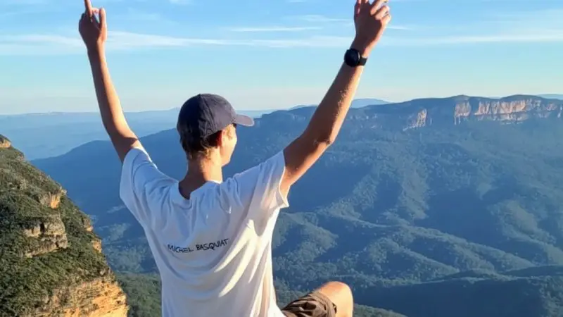 Traveller sits on scenic Blue Mountains cliff edge, arms uplifted, savouring panoramic views during all-inclusive signature day tour.
