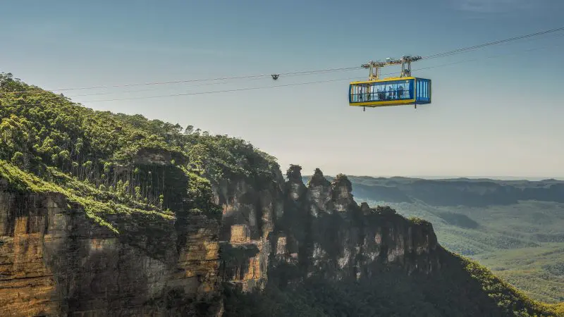 Experience breathtaking views as a vibrant blue and yellow cable car soars above Lyrebird Pass at Scenic World on a sunny day.