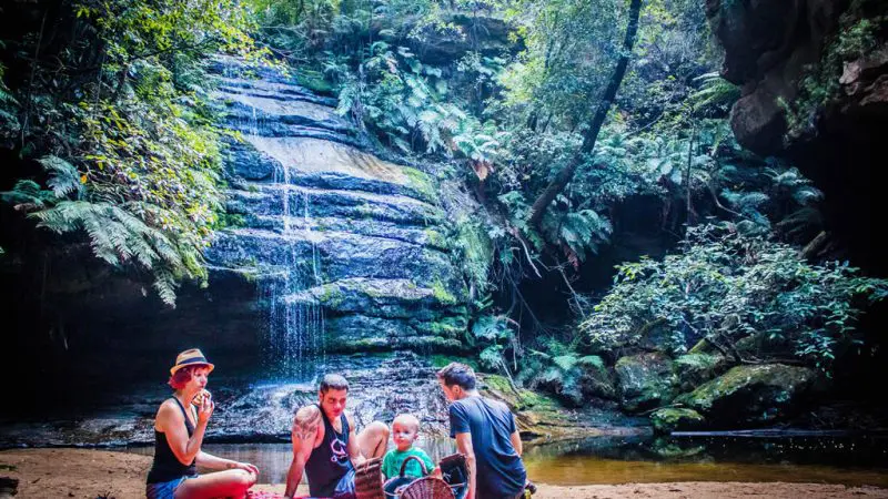 Group of four adults and a child picnic by a scenic forest waterfall after exploring via Explorer Bus Pass Hop-On Hop-Off tour route.