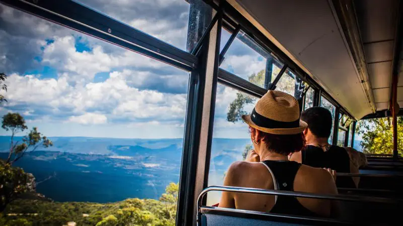 Two travellers with Explorer Bus Passes enjoy stunning mountain scenery through panoramic windows on a sunny Hop On Hop Off tour ride.