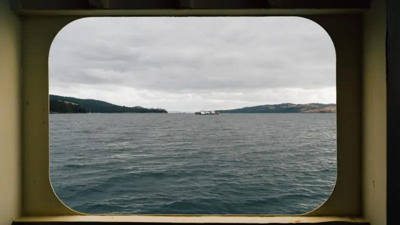 Dramatic cloudy seascape framed by a ship window, showcasing distant Tasmanian land and a tour boat, perfect for Tassie Tours.