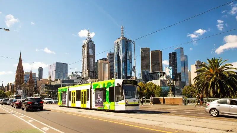 Green and white Melbourne tram travels down a sunlit street with city skyline in the background, highlighting Ultimate Melbourne Adventure.