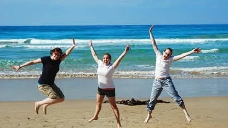 Three people jumping with joy on a sandy beach during the Ultimate 2 Day Great Ocean Road Tour, ocean waves and blue sky in view.