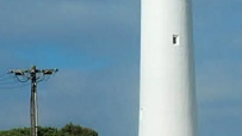 Three tourists stand on a scenic path beside a towering white lighthouse on the Ultimate 2-Day Great Ocean Road Tour in Australia.