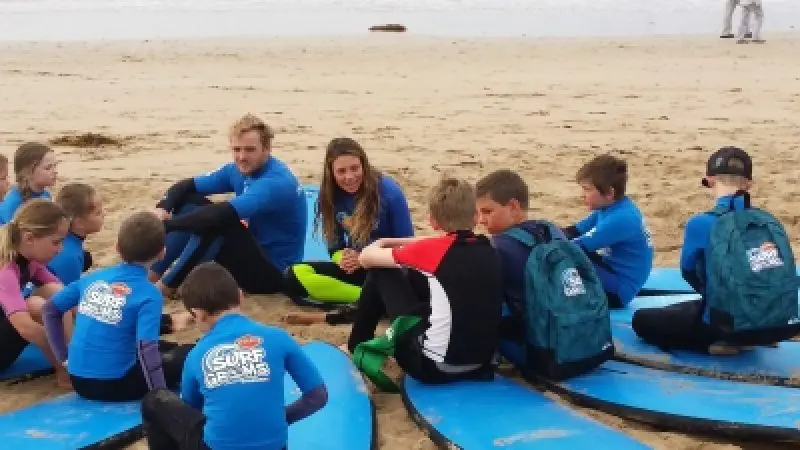 Children and two adults in blue shirts sit on surfboards on a sandy beach, ready for their Learn to Surf Tour surfing lesson.