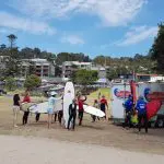 Surf lesson group with surfboards by a Go Ride a Wave trailer in a grassy seaside park during a Learn to Surf Tour.
