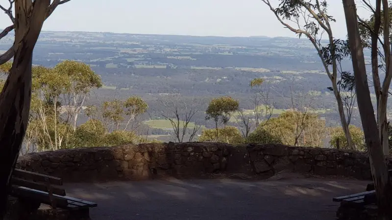 Scenic stone lookout with benches amid lush trees, showcasing panoramic Mt Macedon Ranges and Daylesford views under clear skies.