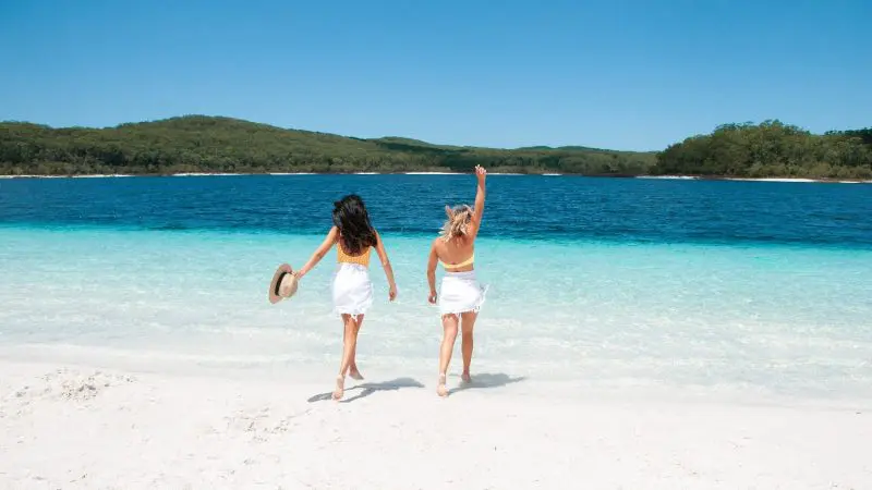 Two women in swimsuits stroll along pristine sand towards crystal-clear blue water on a K'gari Explorer Day Tour departing Rainbow Beach.
