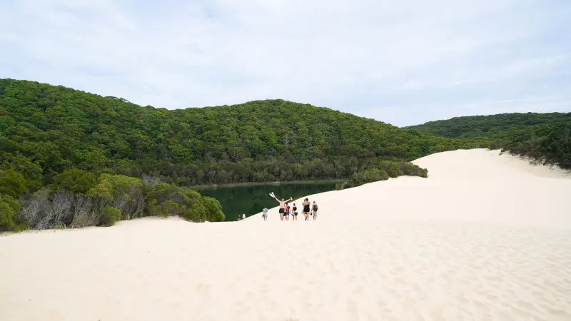 Tour group explores pristine white sand dunes with Island Expeditions K'gari Tours, approaching lush green lake under dramatic clouds.
