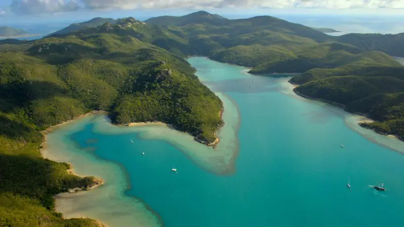Stunning aerial view of turquoise Hook Island bay, boats and Lady Enid sailing beneath scenic partly cloudy Whitsundays sky.
