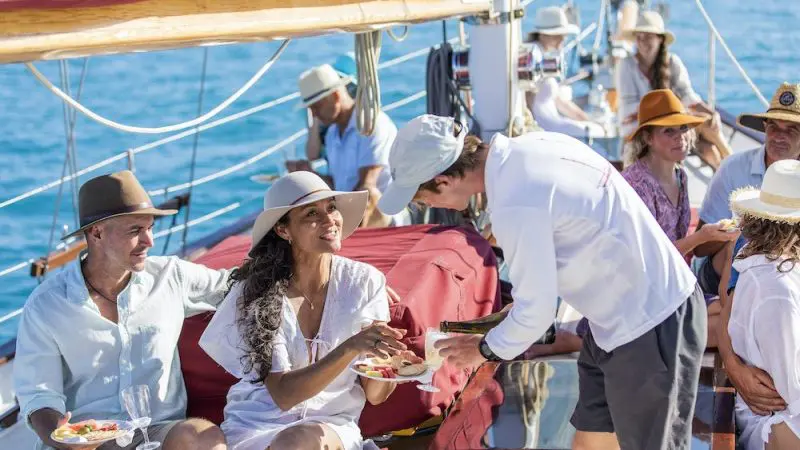 Guests savouring gourmet food and refreshing drinks aboard Lady Enid Sailing, dressed in summer attire, with azure ocean and Hook Island.