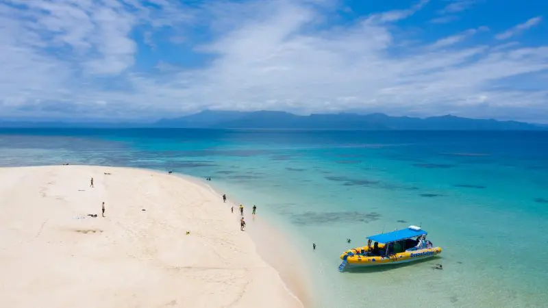 Yellow boat anchored by pristine white sand beach, promising Great Barrier Reef Eco Adventure amid crystal-clear waters and mountain views.