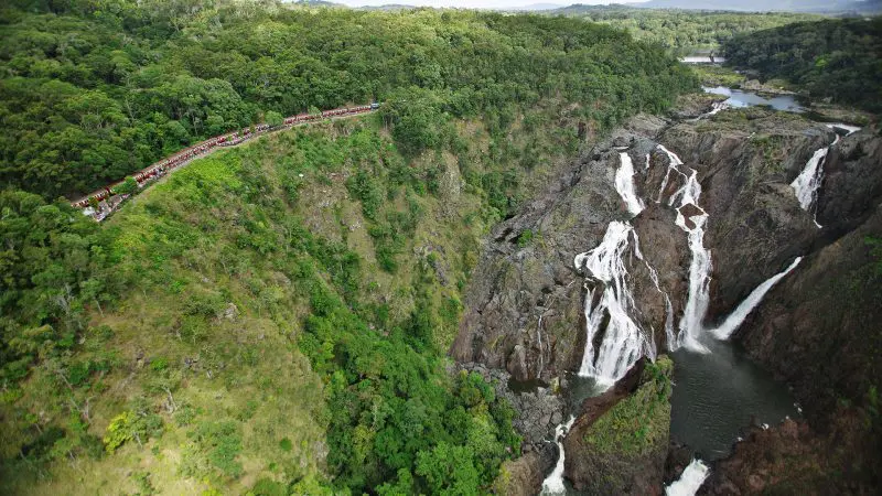 Stunning aerial shot of the iconic Kuranda Train curving through vibrant rainforest beside a waterfall, featured in Skyrail adventure tours.