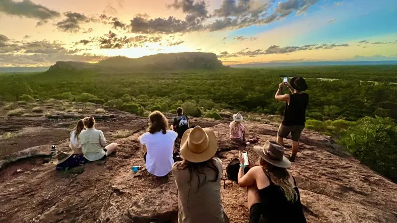 Adventurers atop a rocky hilltop watch a stunning Kakadu sunset after a 1 Day Wilderness Escape and thrilling Crocodile Cruise.