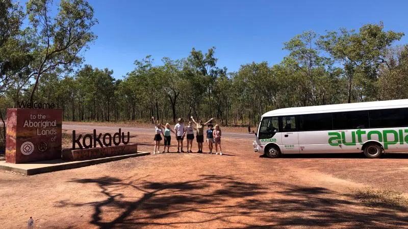 Tourists gather by the Kakadu National Park sign and tour coach for their 1 Day Kakadu Wilderness Escape with Crocodile Cruise adventure.