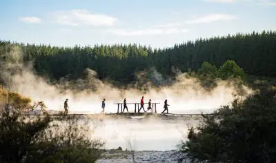 Six people cross a scenic bridge over steaming water; lush trees and greenery surround them beneath a vibrant blue sky.