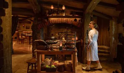 A woman in a white dress stands in a warm, rustic kitchen with exposed wooden beams, brick oven, fresh foods and ingredients displayed.