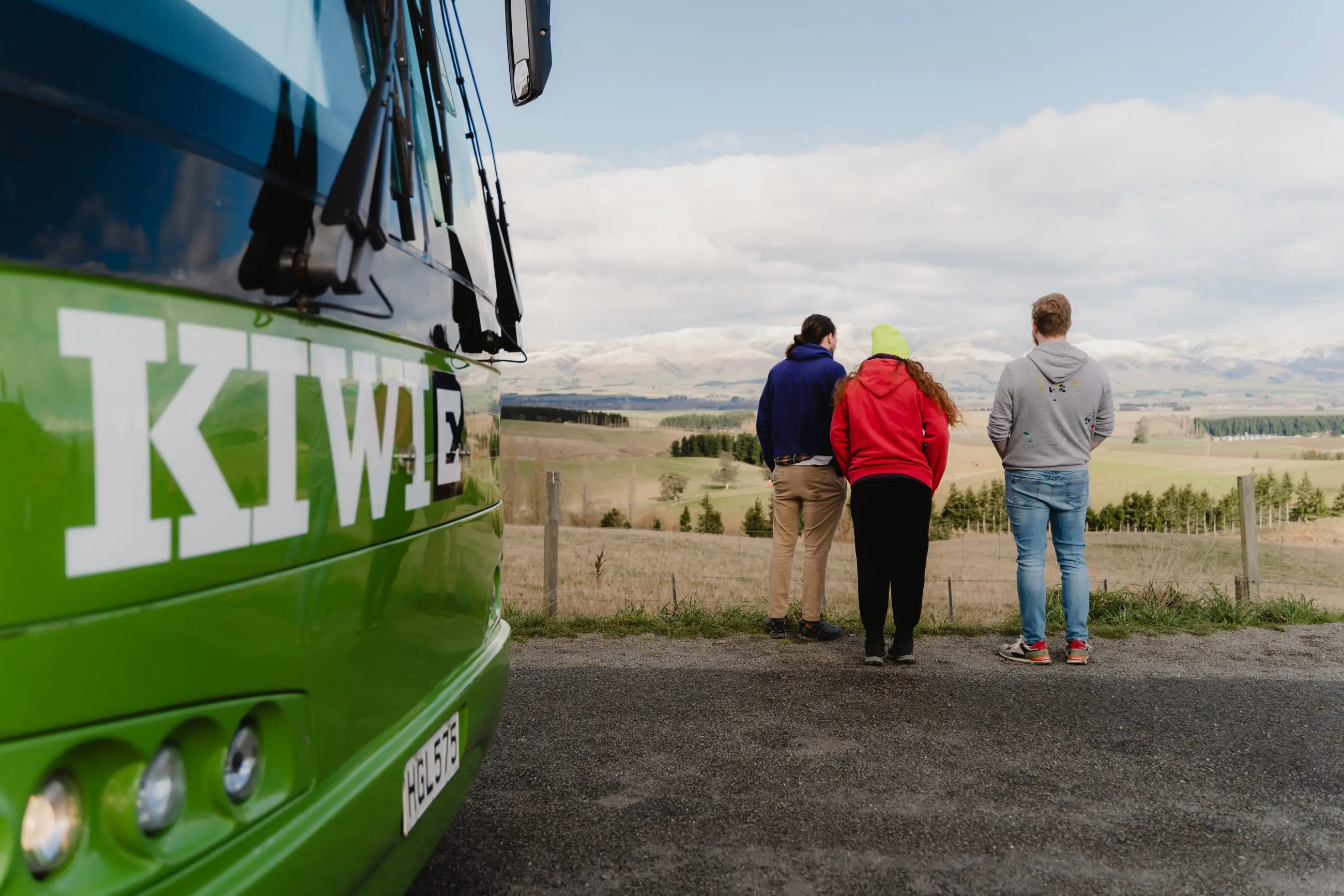 A diverse group of people stands on a paved road beside a vibrant green coach, ready for travel or tours, under clear daylight.