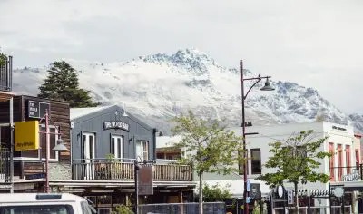 Vibrant city centre street lined with shops and cafés, scenic snowy mountain backdrop, lush trees, and parked cars in the foreground.